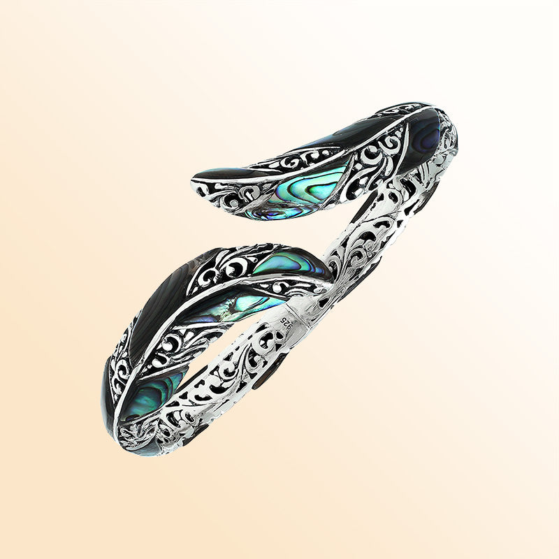 Handcrafted sterling silver feather-inspired bangle featuring luminous Abalone Shell inlays, ornate Balinese scroll engravings, and a secure hinge lock for expressive, artistic everyday wear.