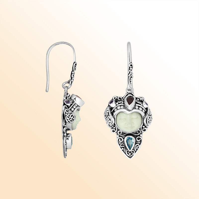 Sterling silver artisan earrings featuring a serene Bone Face carving paired with Blue Topaz Quartz and Garnet Quartz stones, surrounded by ornate Balinese scrollwork for expressive personal styling.